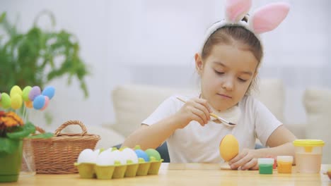 Little-playful-girl,-wearing-bunny-ears-on-her-head-is-choosing-an-a-red-colour-to-paint-an-egg-and-calmly-is-painting-an-Easter-egg.-Girol-has-painted-an-heart-on-it,-then-shows-a-gesture-of-love.