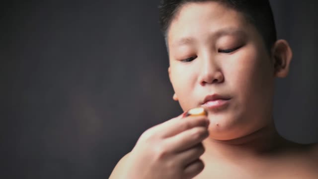 Großer-asiatischer-Vorzehn-Junge,-der-Donut-isst,-während-er-seinen-Bauch-berührt,-Zeitlupe-bei-niedrigem-Licht.