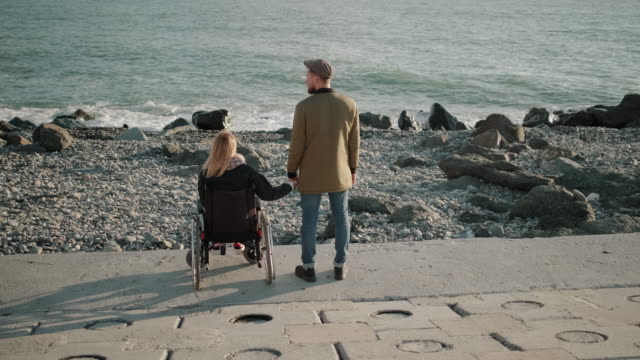 Mujer-en-silla-de-ruedas-usuario-y-hombre-sano-están-cogidos-de-la-mano-y-mirando-el-paisaje-marino