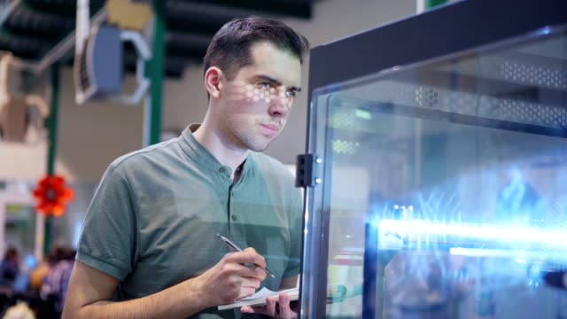 Fokussierter-junger-Techniker,-der-an-der-3D-Druckmaschine-im-Labor-steht,-dreidimensionale-Druckprozesse-beobachtet-und-Notizen-im-Notizblock-macht