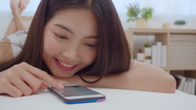 Cámara-lenta---Joven-mujer-asiática-usando-teléfonos-inteligentes-revisando-las-redes-sociales-sintiéndose-feliz-sonriendo-mientras-se-acuesta-en-la-cama-después-de-despertarse-por-la-mañana,-atractiva-chica-japonesa-sonriendo-relajarse-en-el-dormito