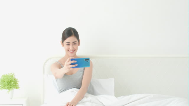 Asiatische-Frau-posting-und-fotografieren-für-ein-neues-Bild-von-sich-mit-Smartphone-im-Bettzimmer.-Konzept-der-Technologie,-Kommunikation,-Webcam,-hochgeladen-und-Selfie.