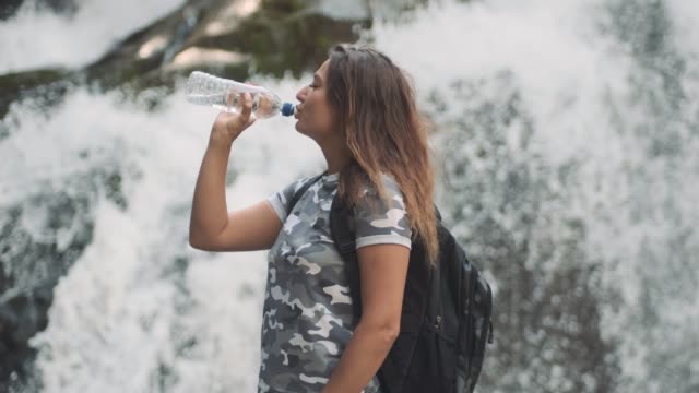 Attraktive-Mädchen-trinkt-kaltes-Wasser-aus-einer-Flasche-und-löscht-Durst,-während-auf-einem-Stein-in-der-Nähe-eines-Wasserfalls-stehen