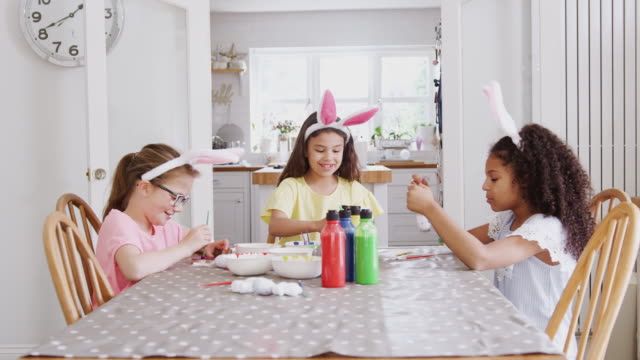 Grupo-de-chicas-sentadas-alrededor-de-la-mesa-de-la-cocina-con-orejas-de-conejo-decorando-huevos-para-Semana-Santa---tiro-en-cámara-lenta