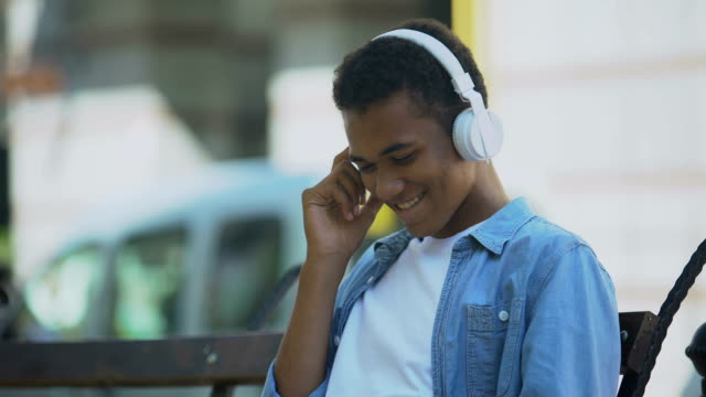 Positiver-junger-Mann,-der-Musik-in-Kopfhörern-hört-und-auf-der-Bank-sitzt