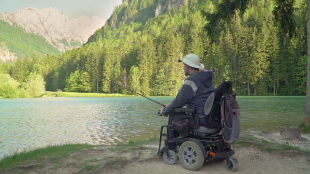 Zeitlupe-von-behinderten-Fischer-in-einem-elektrischen-Rollstuhl-Angeln-in-schönen-See-in-der-Nähe-von-Wald-und-Berg-im-Rücken