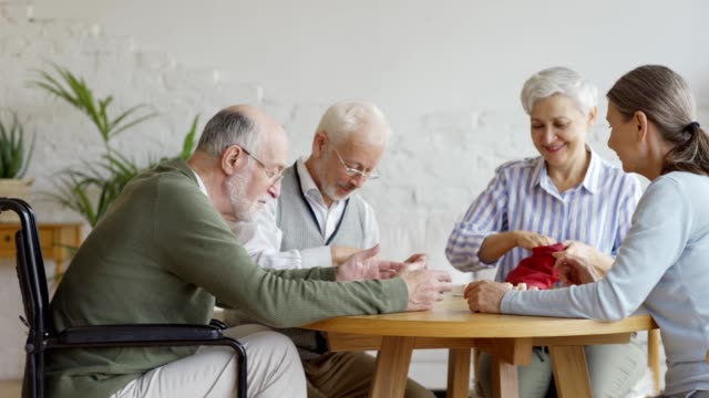 Tracking-Aufnahme-von-Gruppe-von-vier-fröhlichen-rentnern-Senioren,-zwei-Männer-und-zwei-Frauen,-Spaß-am-Tisch-sitzen-und-Spielen-Bingo-Spiel-zusammen-in-Pflegeheim