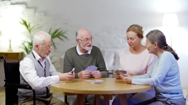 Group-of-four-senior-people,-two-men-including-disabled-one-on-wheelchair-and-two-women,-playing-cards-and-talking-sitting-at-table-in-nursing-home
