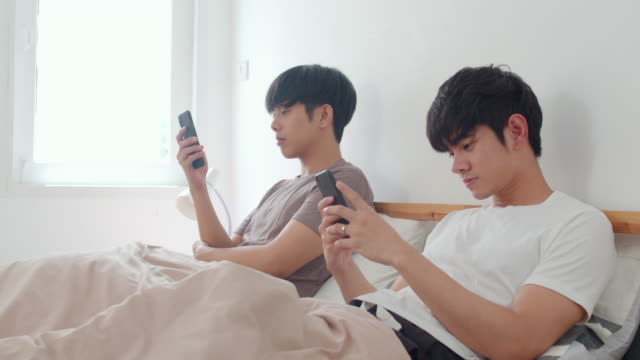 Asian-Gay-couple-using-mobile-phone-at-home.-Young-Asian-LGBTQ-men-happy-relax-rest-together-after-wake-up,-check-social-media-lying-on-bed-in-bedroom-at-home-in-the-morning-concept.-Slow-motion-Shot.