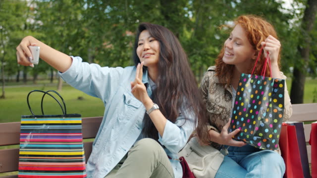 Fröhliche-Mädchen-machen-Selfie-mit-Einkaufstaschen-sitzen-auf-der-Bank-im-Park-posiert