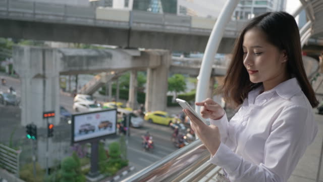 Junge-lächelnde-Geschäftsasiatische-Frau-mit-Smartphone-und-zu-Fuß-auf-der-Stadt-Straße,-während-das-Gespräch-mit-ihrem-Freund-auf-dem-Handy-am-Abend-von-Bangkok-Thailand.
