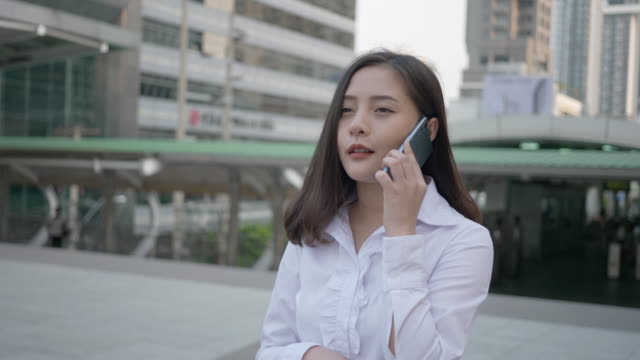 Joven-mujer-asiática-sonriente-usando-el-teléfono-inteligente-en-la-calle-de-la-ciudad-mientras-habla-con-su-amigo-en-el-teléfono-móvil-en-la-noche-de-Bangkok-Tailandia.