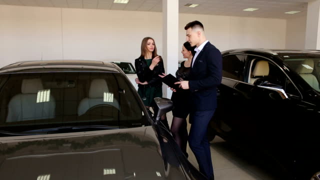 Zwei-Mädchen-gehen-mit-dem-Verkäufer-in-den-großen-Autoshop-und-wählen-ein-neues-Auto.