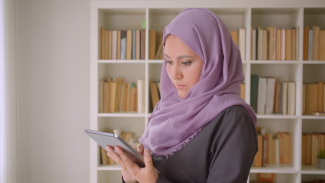 Retrato-de-primer-plano-de-joven-musulmana-en-hiyab-usando-la-tableta-y-mirando-la-cámara-de-pie-en-la-biblioteca-en-el-interior