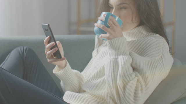 Beautiful-Young-Woman-Using-Smartphone,-Drinks-Tea-while-Sitting-on-the-Chair.-Sensual-Girl-Wearing-Sweater-Surfs-Internet,-Posts-On-Social-Media,-Shares-Pictures-while-Relaxing-in-Cozy-Apartment