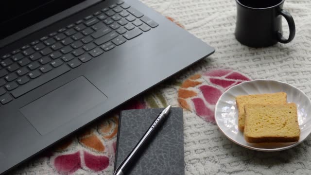 Breakfast-coffee-in-morning-sunlight-with-laptop-computer-black-color-pen-and-personal-organizer-notebook,-ceramic-cup-saucer-and-biscuit-on-top-office-place-working-desk-background.-Lifestyle-image.