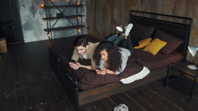Couple-moving-to-new-apartment.-Male-and-female-using-smartphones,-lying-together-on-bed-in-room-with-modern-interior-and-cardboard-boxes