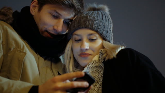 Young-couple-watching-photos-on-smartphone-at-night-in-a-city-street.