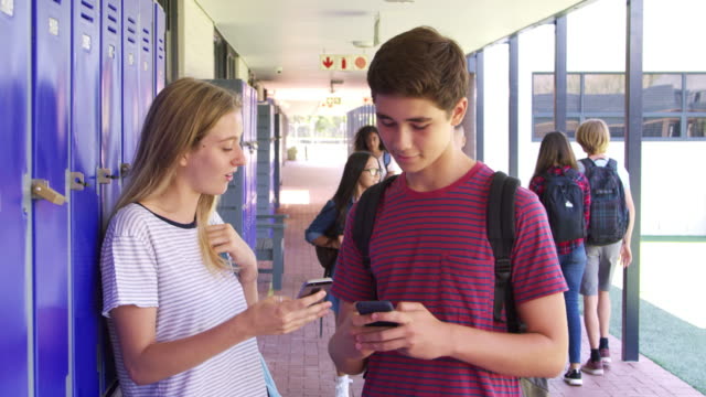 Two-teenage-friends-talking-use-phones-in-school-corridor