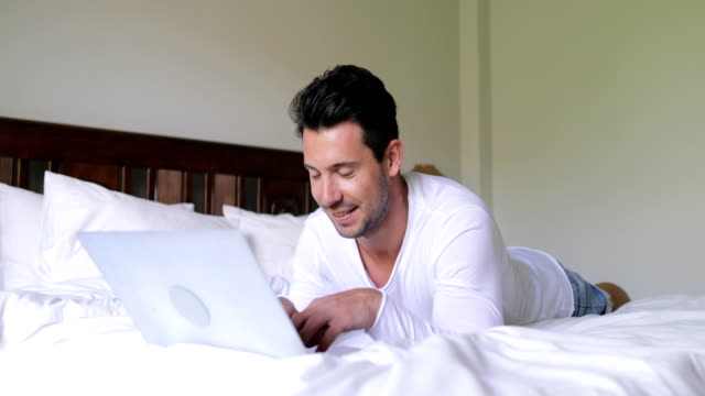 Junger-Mann-mit-Laptop-Computer-am-Bett-glücklich-lächelnd-Kerl-im-Chat-Online,-Schlafzimmer-morgens-liegen