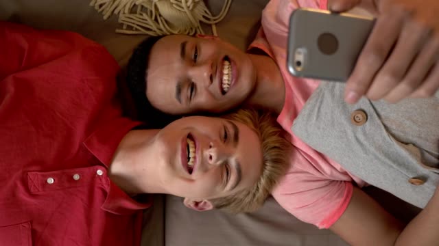 Happy-international-gay-couple-is-lying-on-the-couch-and-making-selfies-on-a-smartphone.-Homeliness,-LGBT-lovers,-happy-gay-family-concept,-laughing.-60-fps