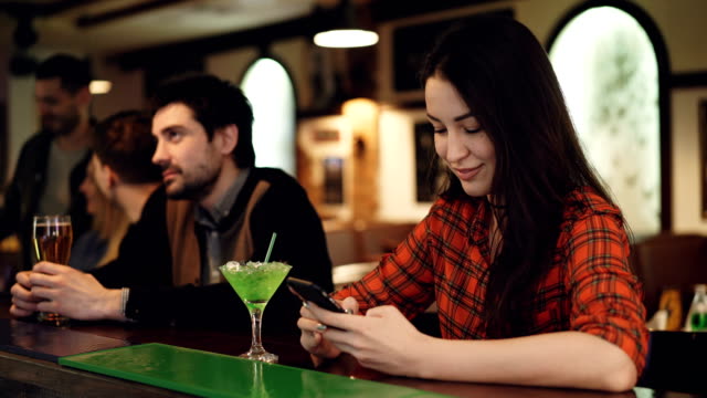 Junge-Schülerin-nutzt-Smartphone-sitzen-in-schicke-Bar-mit-Cocktails.-Sie-ist-Bildschirm-berühren-und-lächelnd.-Moderne-Wege-der-Kommunikationskonzept.