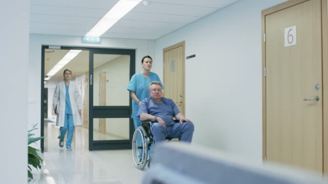 En-el-pasillo-del-Hospital,-la-enfermera-empuja-Senior-hombre-en-silla-de-ruedas,-pacientes-esperan-a-su-médico,-personal-profesional-ocupado-a-pie.-Limpio,-nuevo-Hospital-con-personal-médico-profesional.