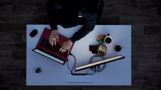 Joven-negro-con-capucha-hacking-laptop-durante-la-noche,-sentado-detrás-del-escritorio-con-ordenador-portátil-y-el-monitor-superior-tiro