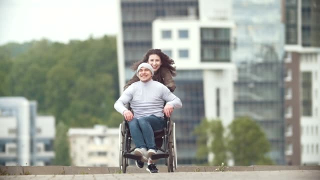 Happy-disabled-man-in-a-wheelchair-with-happy-young-woman-running-at-the-city-street
