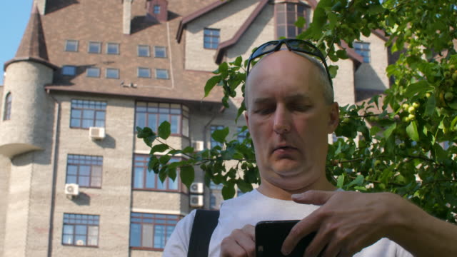 Bald-man-editing-video-on-smartphone-in-city-with-tree-and-building