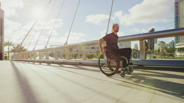 Young-happy-disabled-man-pushing-wheelchair-across-bridge-in-city-with-lens-flare