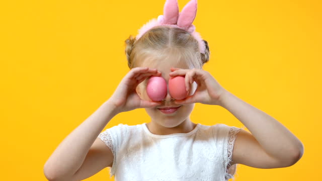 Cute-girl-having-fun-putting-colored-egg-to-eyes-on-yellow-background,-childhood