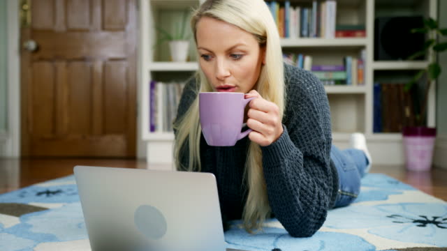 Schöne-blonde-Frau-liegend-auf-Teppich-mit-Laptop-Tee-trinken