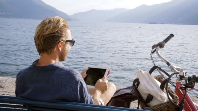 Hombre-joven-con-tableta-digital-en-aldea-Suiza-cerca-de-lago-y-las-montañas-sentado-en-Banco-con-bicicleta-al-lado-de-él.-Concepto-de-personas-viajes-tecnología-dispositivos-portátiles