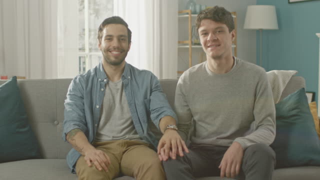 Cute-Attractive-Male-Gay-Couple-Sit-Together-on-a-Sofa-at-Home.-Boyfriend-Puts-His-Hand-on-Partner's.-They-are-Happy-and-Smiling.-They-are-Casually-Dressed-and-Their-Room-Has-Modern-Interior.