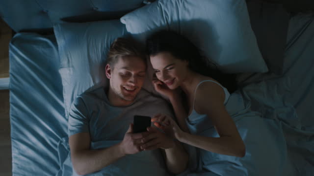 Happy-Young-Couple-Use-Smartphone-in-Bed-at-Night,-They-Share-Screen,-Browsing-Through-Social-Networks,-Sharing-Pictures,-Using-Internet-und-Having-Fun.-Top-View-Zoom-aus-Kameraschuss