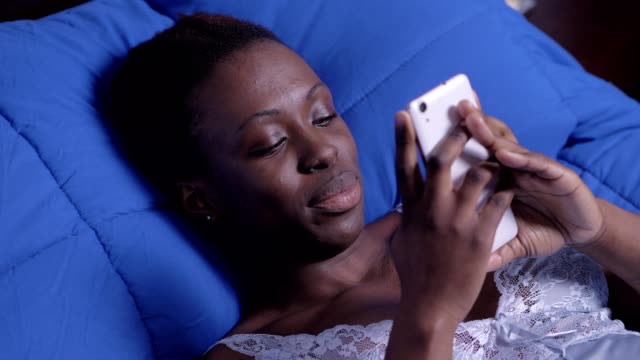 Sonriente-mujer-negro-americano-navegando-red-con-teléfono-celular-en-casa,-acostado-en-la-cama