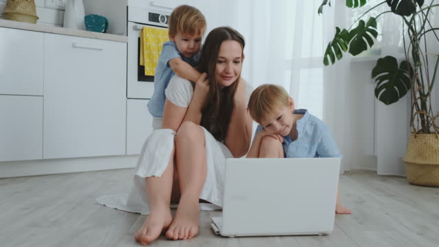 Moderne-Wohnung-liebende-Mutter-und-zwei-kleine-Söhne,-die-auf-dem-Boden-im-Wohnzimmer-sitzen,-schauen-auf-den-Laptop-Bildschirm.-Kinder-mit-Mama-spielen-auf-einem-Laptop