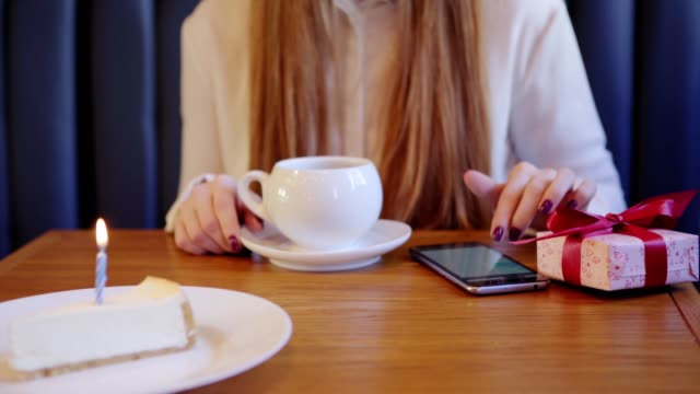 Close-up-of-unrecognizable-girl-browsing-smartphone-while-waiting-for-her-friend-at-table-in-cafe-to-congratulate-on-birthday.-Gift-box-and-cheesecake-with-burning-candle-prepared-for-friend.