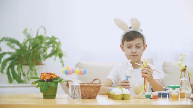 Young-adorable-boy-is-sitting-at-the-table-full-of-Easter-decorations-and-is-playing-with-Easter-bunnies-in-his-hands.-Bunnies'-discussion.-Who-would-paint-an-egg-Bunny-theatre.