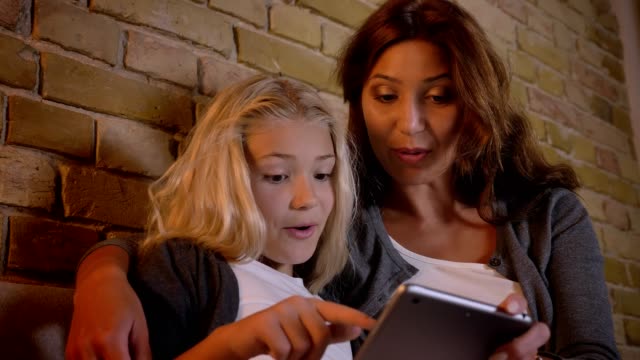 Closeup-shoot-of-young-caucasian-mother-and-her-pretty-small-daughter-using-the-tablet-and-getting-excited-sitting-on-the-couch-indoors-at-home