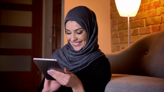 Closeup-portrait-of-adult-attractive-muslim-female-in-hijab-surfing-web-on-the-tablet-and-smiling-while-sitting-on-the-floor-in-doorway-in-a-cozy-apartment