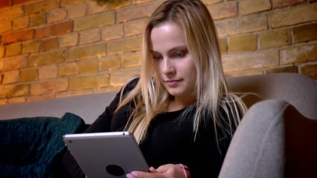 Closeup-portrait-of-young-pretty-blonde-haired-female-using-the-tablet-and-lying-on-the-couch-laidback-indoors-at-cozy-home