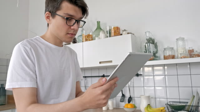 Attractive-man-at-home-using-tablet-in-kitchen-sending-message-on-social-media-smiling-enjoying-modern-lifestyle-wearing-white-shirt