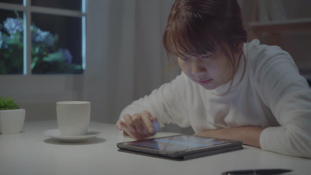 Junge-asiatische-Frau,-die-spät-mit-Tablet-Check-Social-Media,-während-entspannen-Sie-auf-dem-Schreibtisch-in-der-Nacht-im-Wohnzimmer-zu-Hause.-Genießen-Sie-die-Zeit-im-heimischen-Konzept.