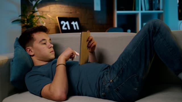 Ein-hübscher-Teenager-liegt-bequem-auf-der-Couch-und-scrollt-das-Futter-im-sozialen-Netzwerk-auf-seinem-Tablet