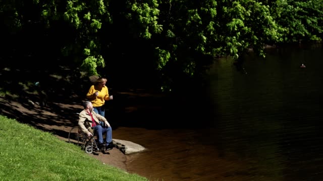 Wife-or-caregiver-bringing-coffee-for-disabled-man-in-wheelchair-waiting-by-lake-in-park.-Couple-having-coffee-and-talking-on-windy-summer-day
