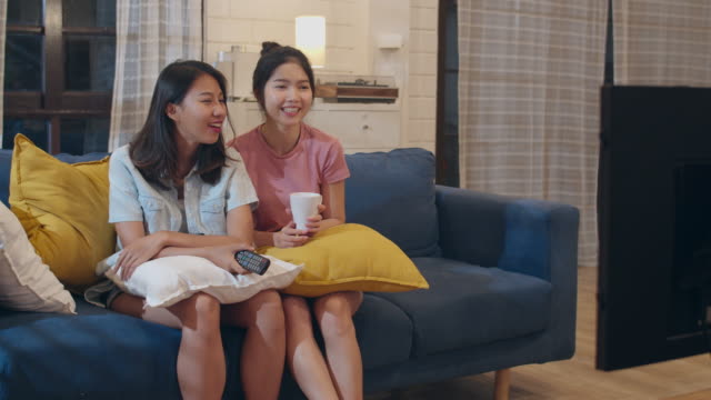 Lesbian-lgbt-women-couple-watching-television-at-home,-Asian-female-lover-feeling-happy-funny-moment-looking-drama-entertainment-together-on-sofa-in-living-room-in-night-concept.