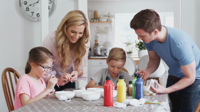 Familie-sitzt-um-Küchentisch-mit-Eltern,-die-Kindern-helfen,-Eier-für-Ostern-zu-malen---in-Zeitlupe-gedreht