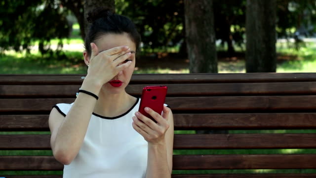 Nahaufnahme-Porträt-Ein-schönes-junges-Mädchen-in-einem-weißen-T-Shirt-plaudert-in-sozialen-Netzwerken-auf-ihrem-Smartphone,-während-sie-auf-einer-Bank-in-einem-Park-sitzt.